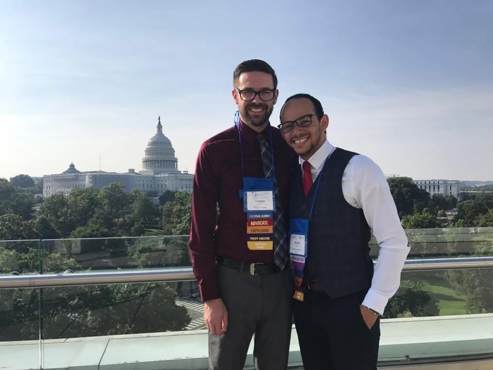 Two smiling men wearing conference lanyards pose together on an outdoor rooftop terrace or balcony. One man wears a maroon dress shirt and tie, and the other wears a dress shirt, red tie, and vest. The United States Capitol dome is visible in the background against a blue sky.