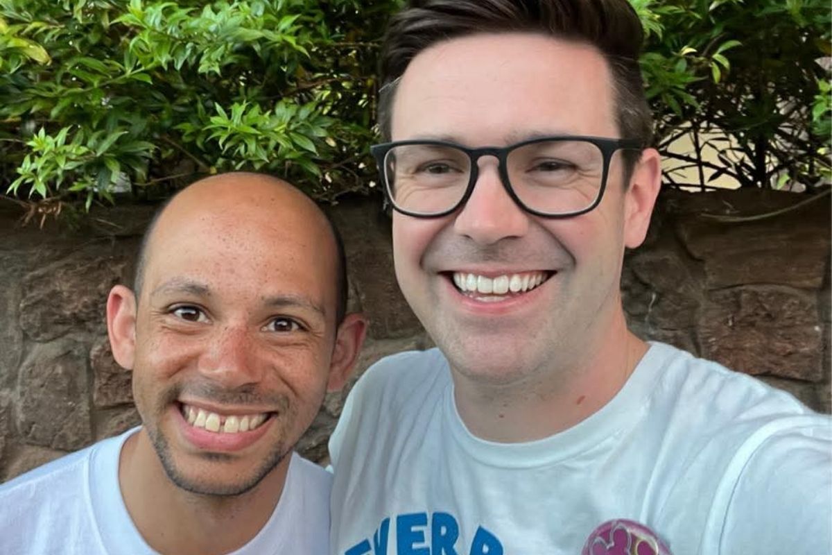 A close-up selfie of two smiling men wearing white t-shirts. The man on the left is bald with a light beard, and the man on the right wears thick black glasses. They are standing outdoors in front of a stone wall and green leafy bushes.