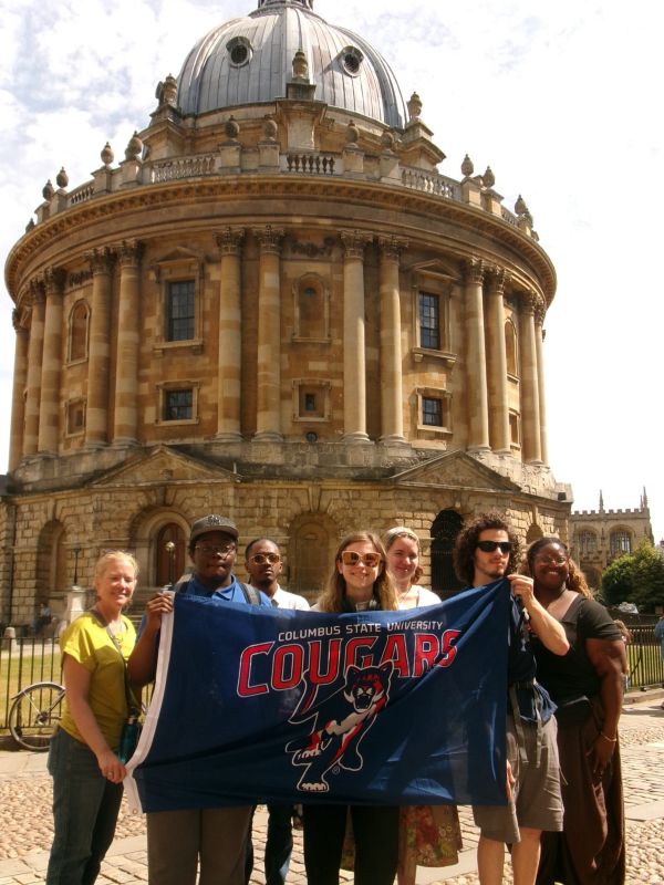 A group of seven people holding up a large blue Columbus State University Cougars mascot banner. They are posing outdoors on a cobblestone plaza directly in front of the historic, circular Radcliffe Camera building in Oxford, England.