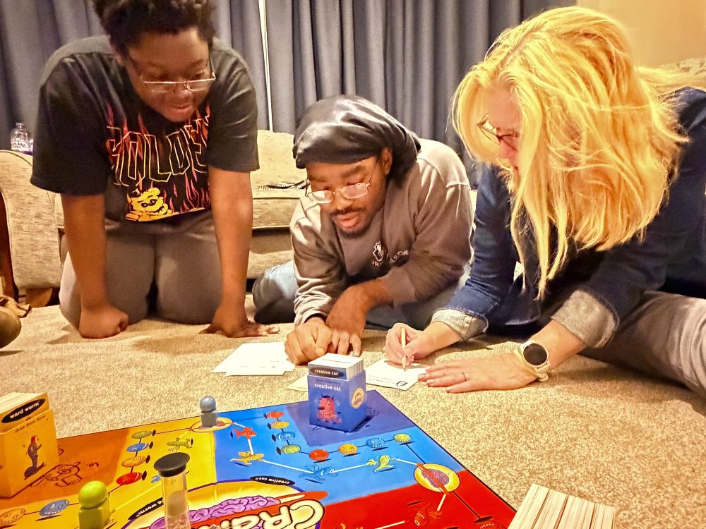 Three people sitting on a carpeted floor playing the board game Cranium. Two students watch closely as a blonde woman in a denim shirt writes on a small piece of paper. The colorful game board, an hourglass timer, game pieces, and a deck of blue cards are spread out in the foreground.