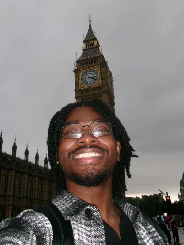 A close-up selfie of a smiling young man with glasses and dreadlocks looking up at the camera. In the background, the Elizabeth Tower (Big Ben) and a portion of the Palace of Westminster rise against an overcast, grey sky.
