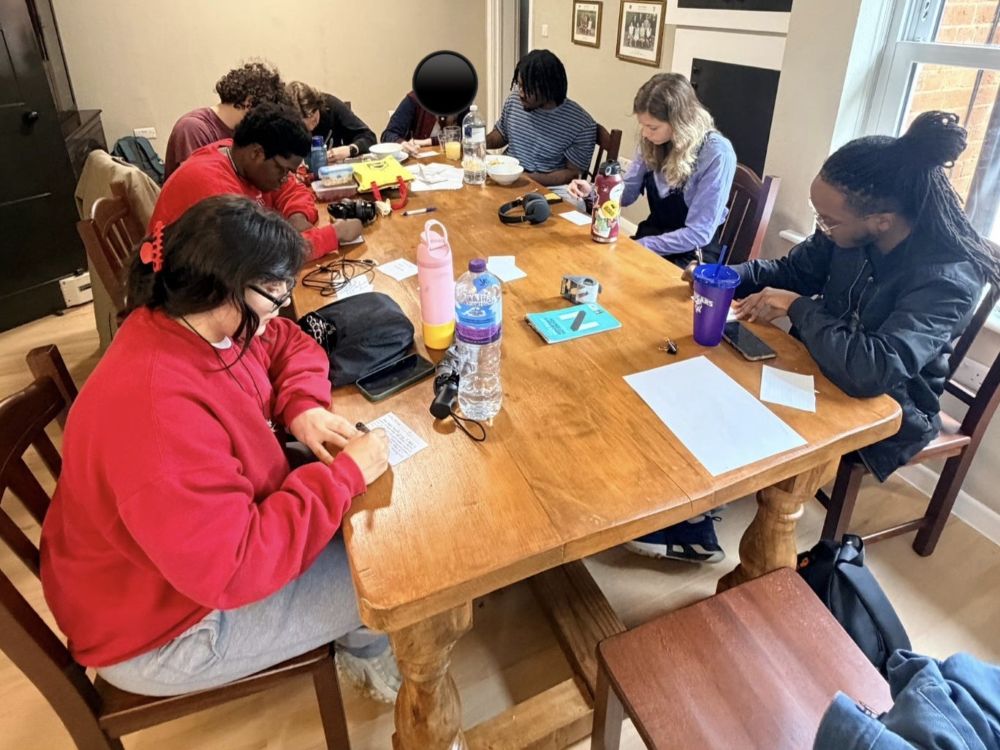 A group of students sitting around a large, rectangular wooden dining table in a brightly lit room with light wood floors. They are focused on individual tasks, with several members actively writing on papers. The table is scattered with personal items, including water bottles, pens, electronics, and notebooks.