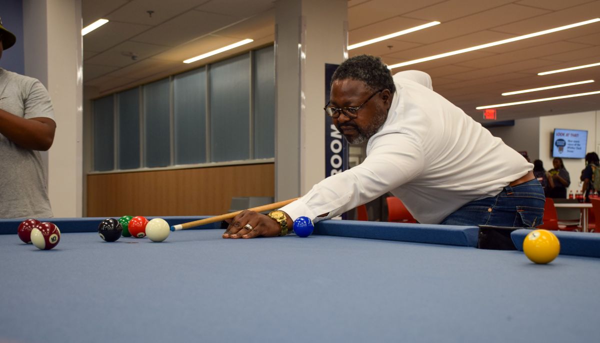 A man in a white dress shirt lines up a shot on a blue pool table during a ribbon-cutting event in a modern indoor student lounge or recreation room.