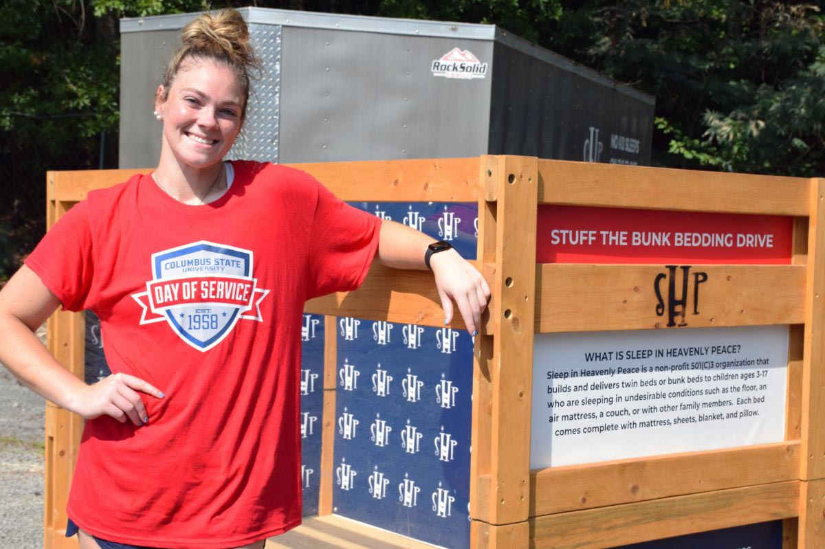 A smiling young woman wearing a red "Columbus State University Day of Service" t-shirt rests her arm on a wooden bunk bed for the "Stuff the Bunk Bedding Drive," hosted by the nonprofit organization Sleep in Heavenly Peace.