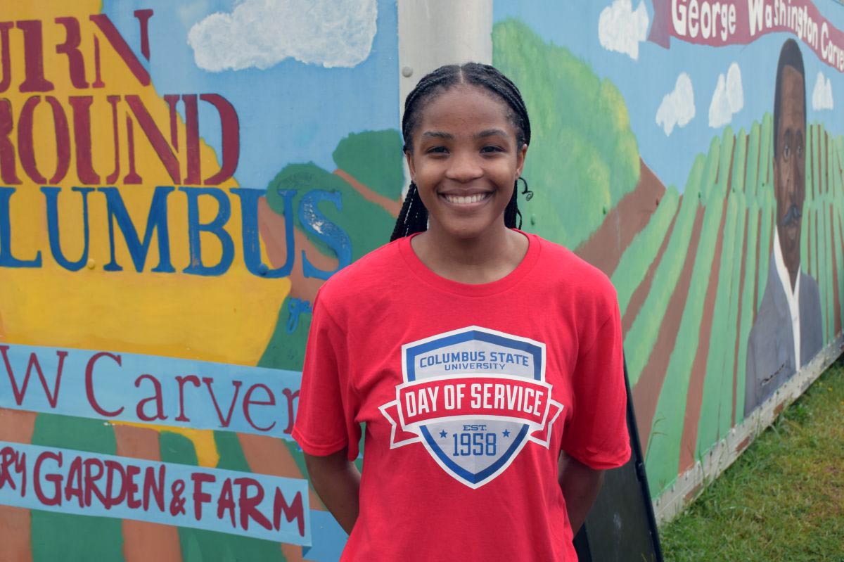 A smiling young woman in a red "Columbus State University Day of Service" t-shirt stands outdoors in front of a colorful painted mural. The mural reads "Turn Around Columbus" and "George Washington Carver Victory Garden & Farm" alongside a portrait of George Washington Carver.