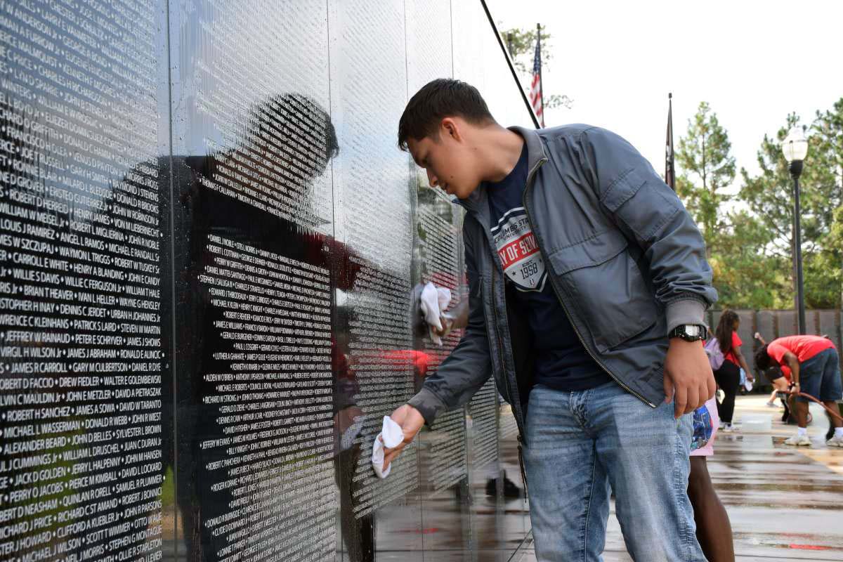 A young man in a gray jacket and a Columbus State University Day of Service t-shirt leans forward to wipe down the polished black granite panels of a memorial wall filled with engraved names.