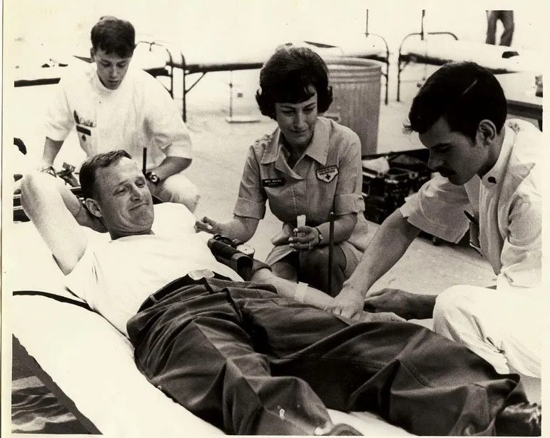 A vintage black-and-white photograph showing a blood drive setup, with Hal Moore lying on a cot as a Red Cross staff and Julie Moore assist.