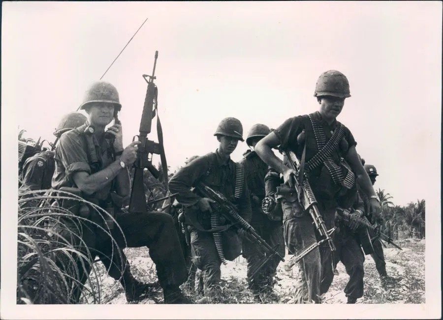 A historical black-and-white photograph depicting soldiers on field operations during the Vietnam War era, featuring a radioman coordinating communications near concertina wire.