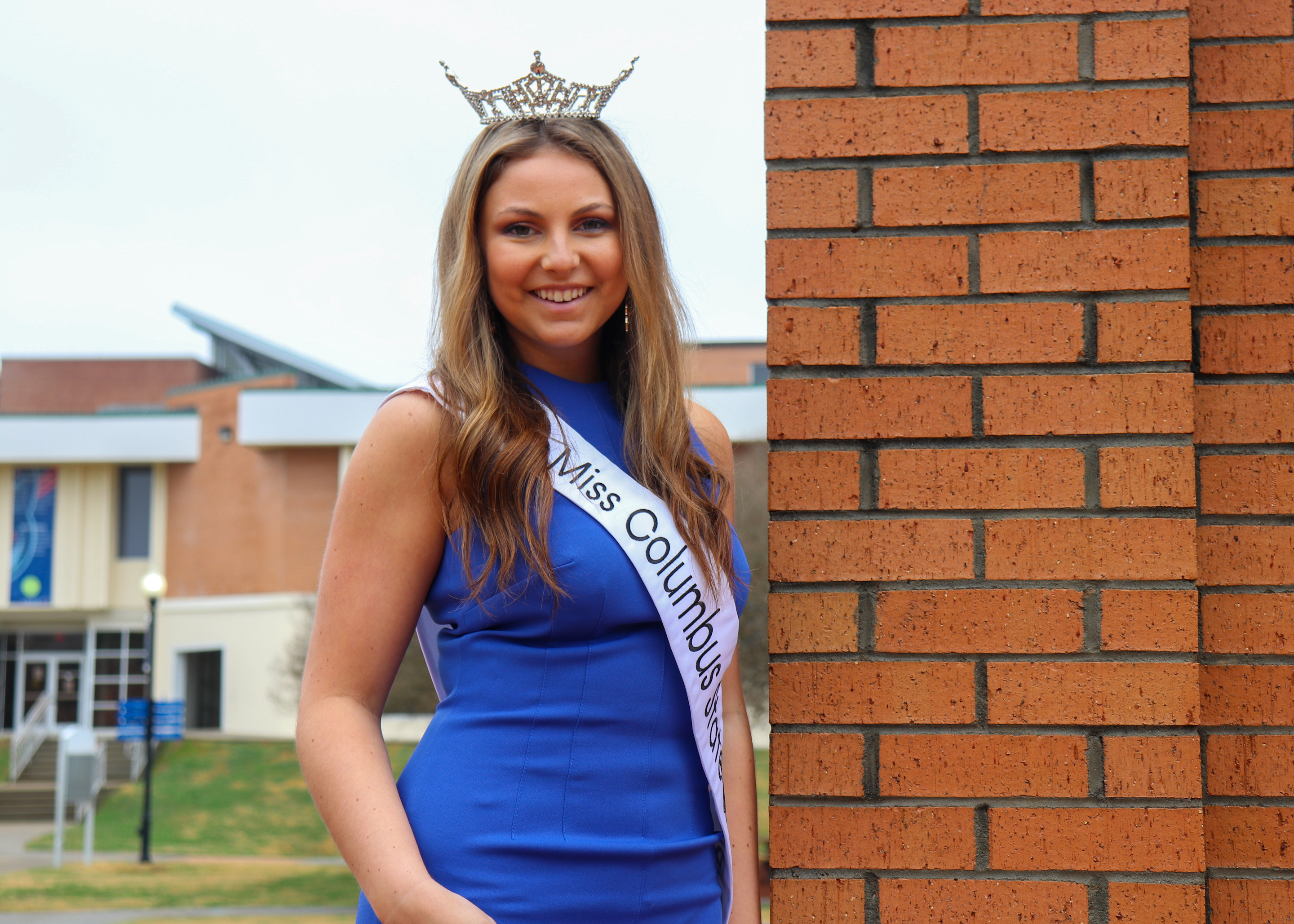 Miss CSU 2022 Tori Svenson in blue dress, crown and sash stands next to brick wall at clocktower with Davidson center behind her.