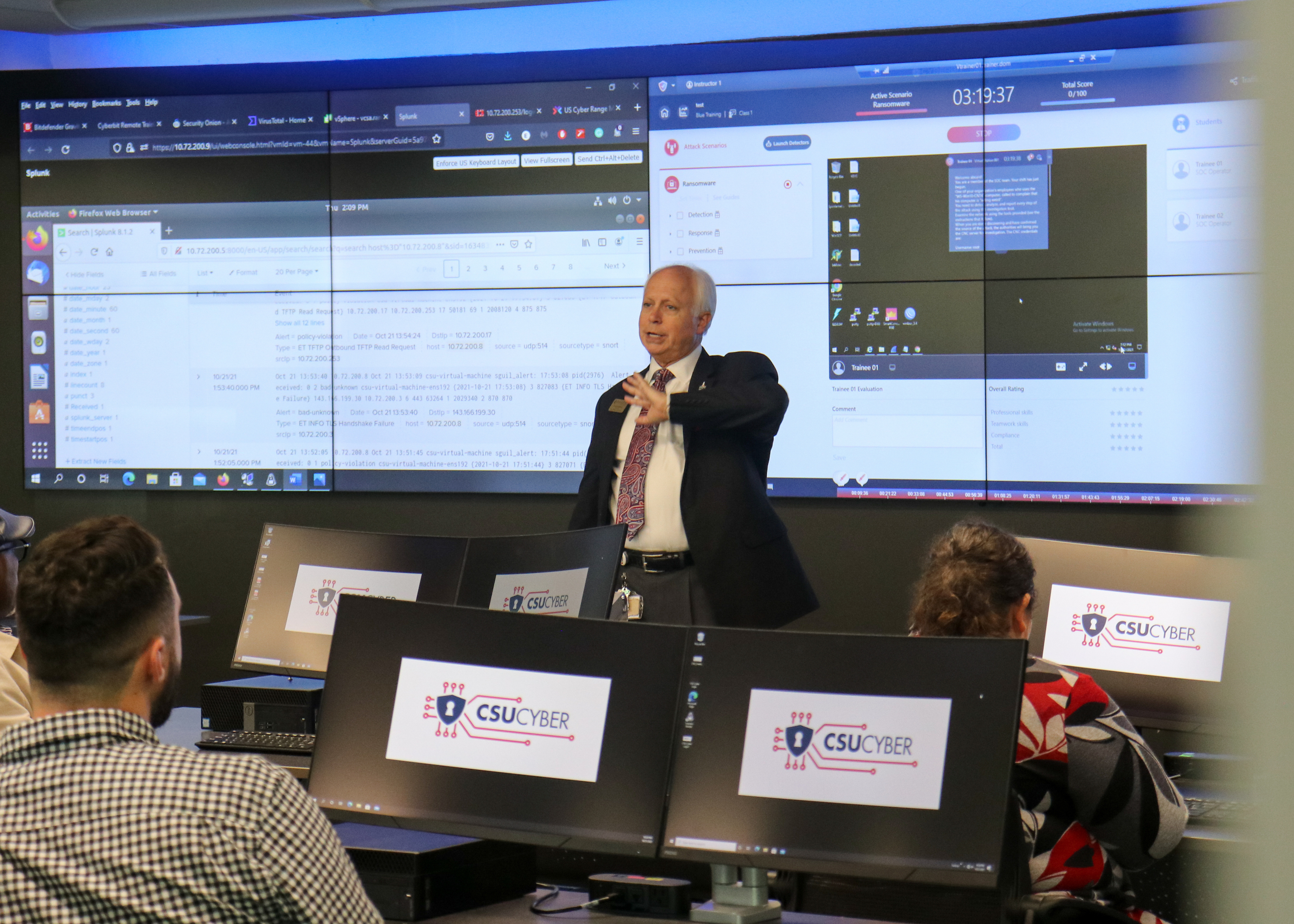A man in a suit stands in front of a computer lab talking to attendees. Behind him are large wall monitors with cyber language and coding on them.
