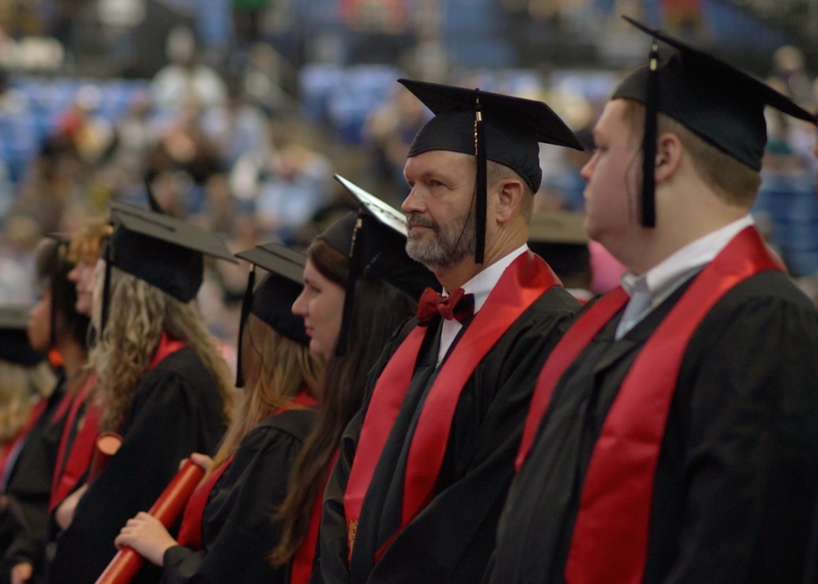 Jim Money in black gown with red stole and grad cap stands in line of graduates facing forward, looking to the left of the picture.