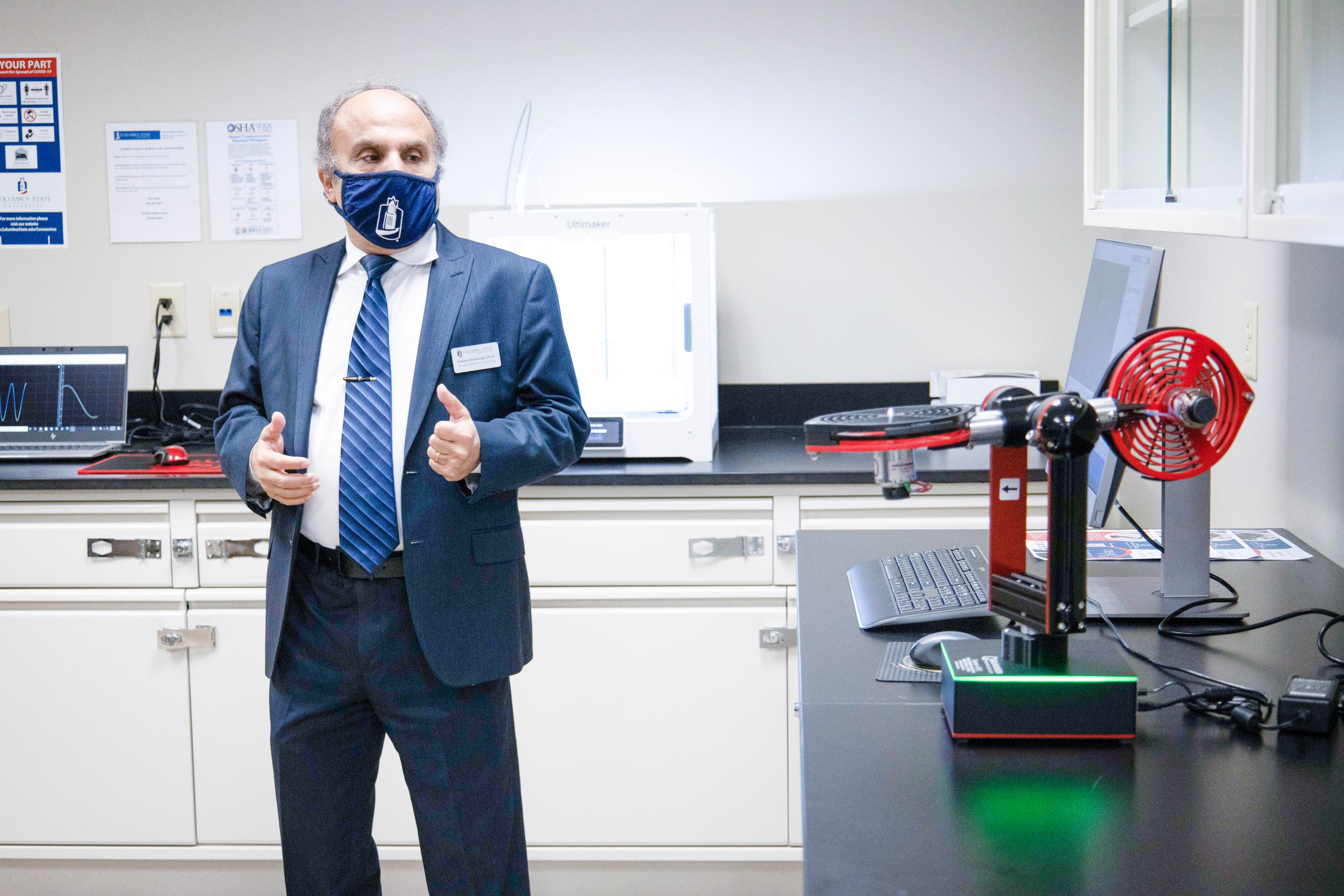 Dr. Reyhanoglu in a suit stands near a red and black robot on a laboratory desk.