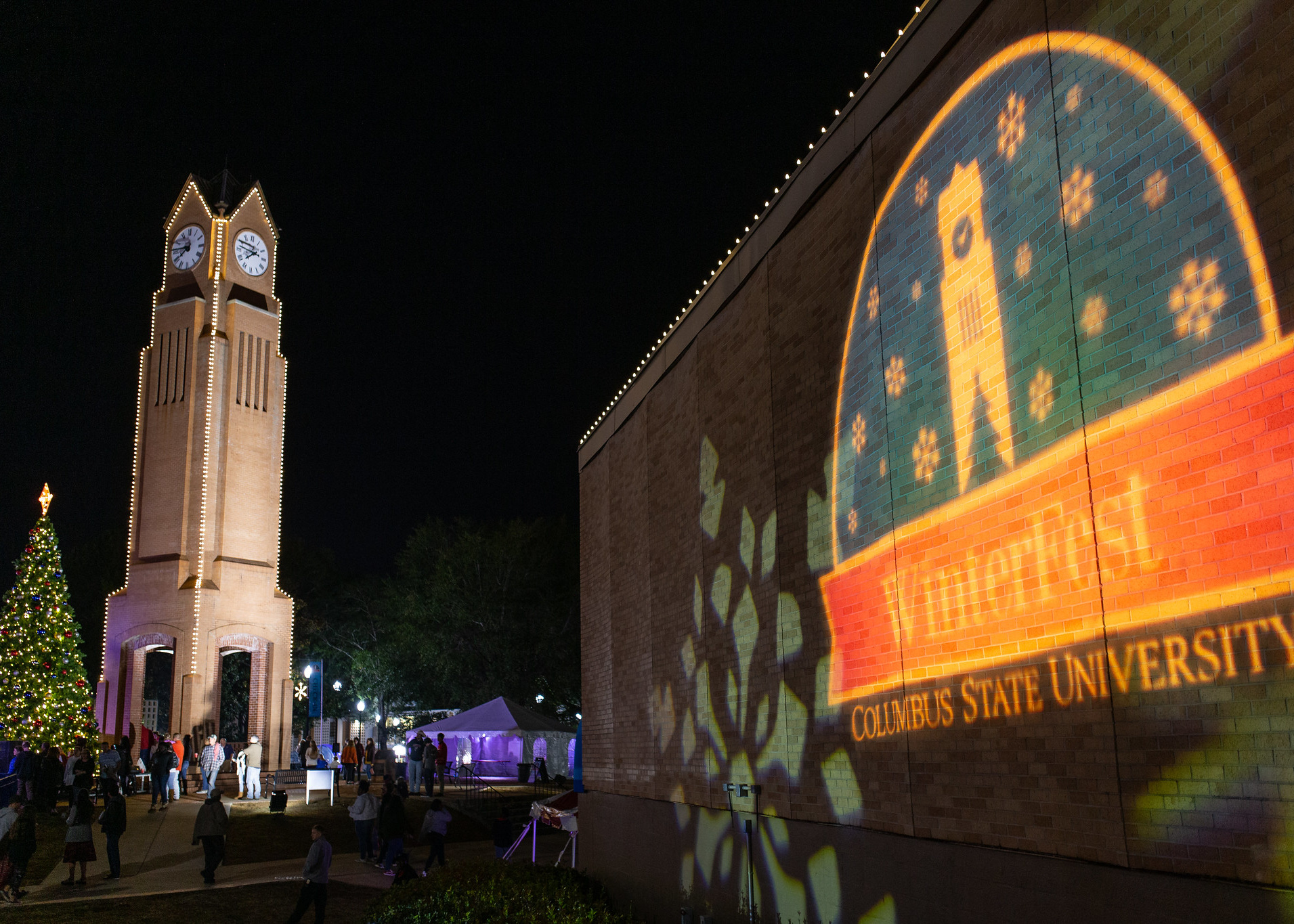 CSU clocktower is lit up next to a tall Christmas tree in the left background. In the right foreground, the Winterfest snow globe logo is reflected on a brick building.