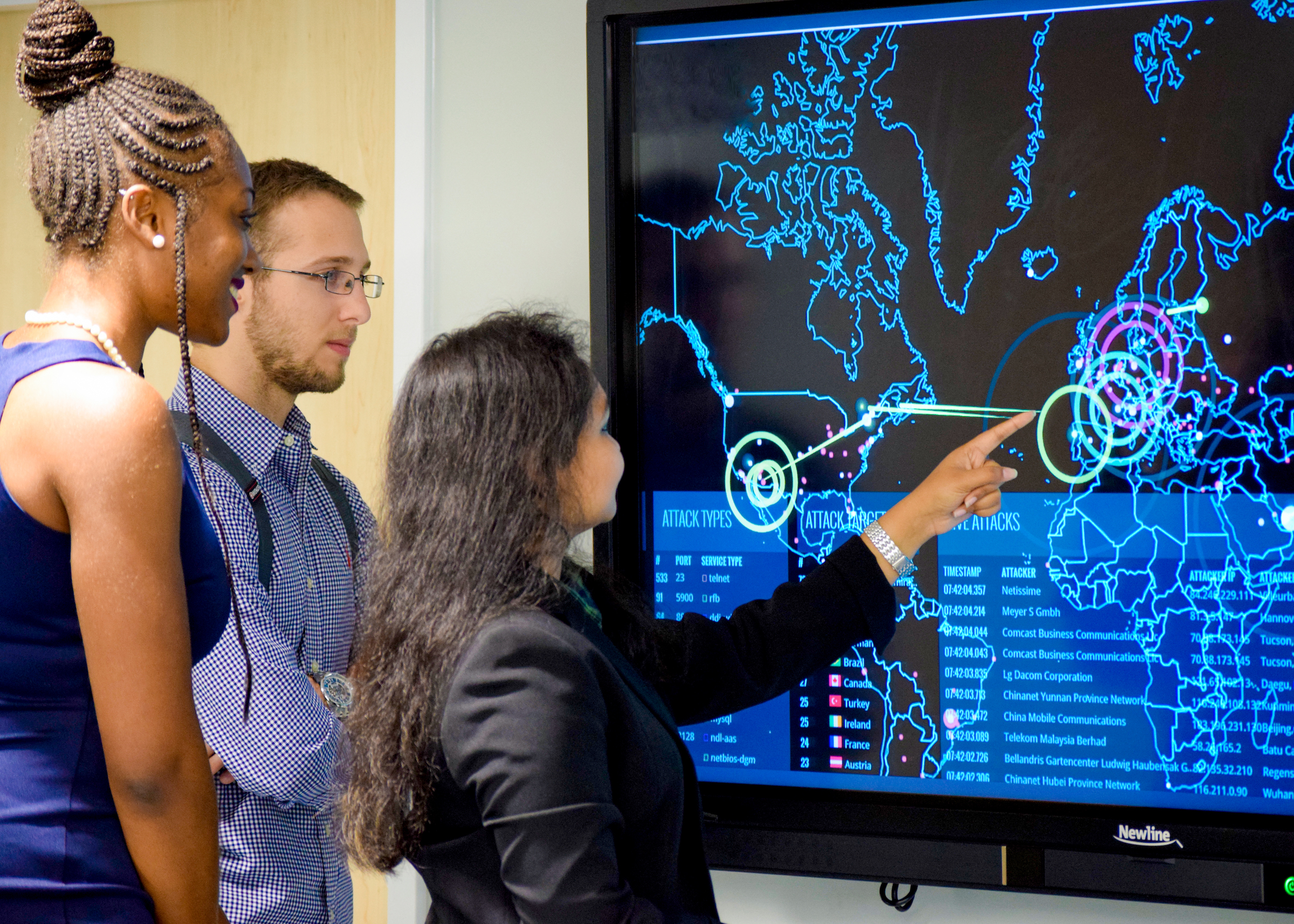 Three students stand next to a large monitor showing a cyber attack map of the world. One student is pointing to the map.
