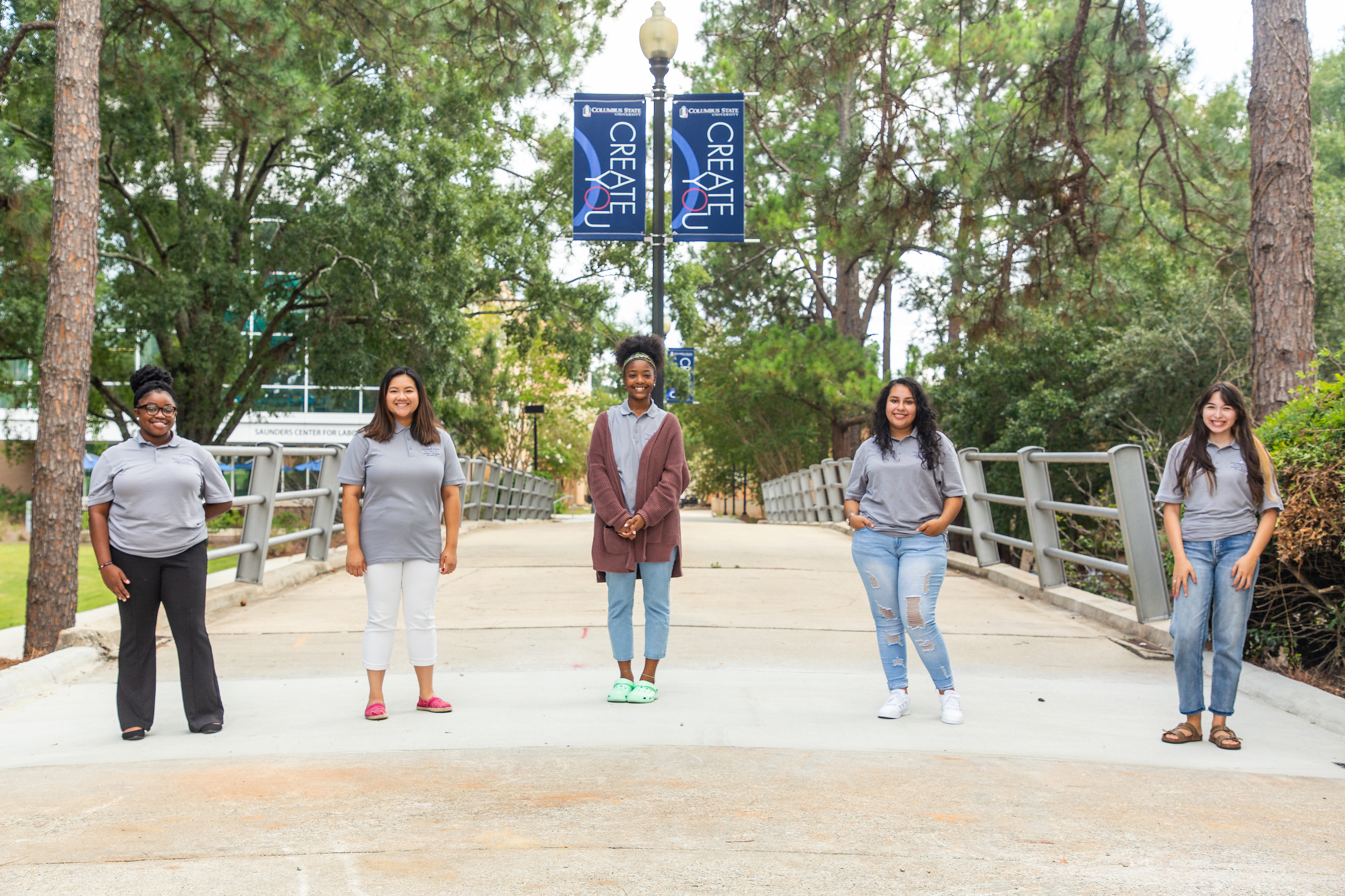 Five female students stand spread apart on a bridge walkway with the CSU Create You banners on a lightest behind them.