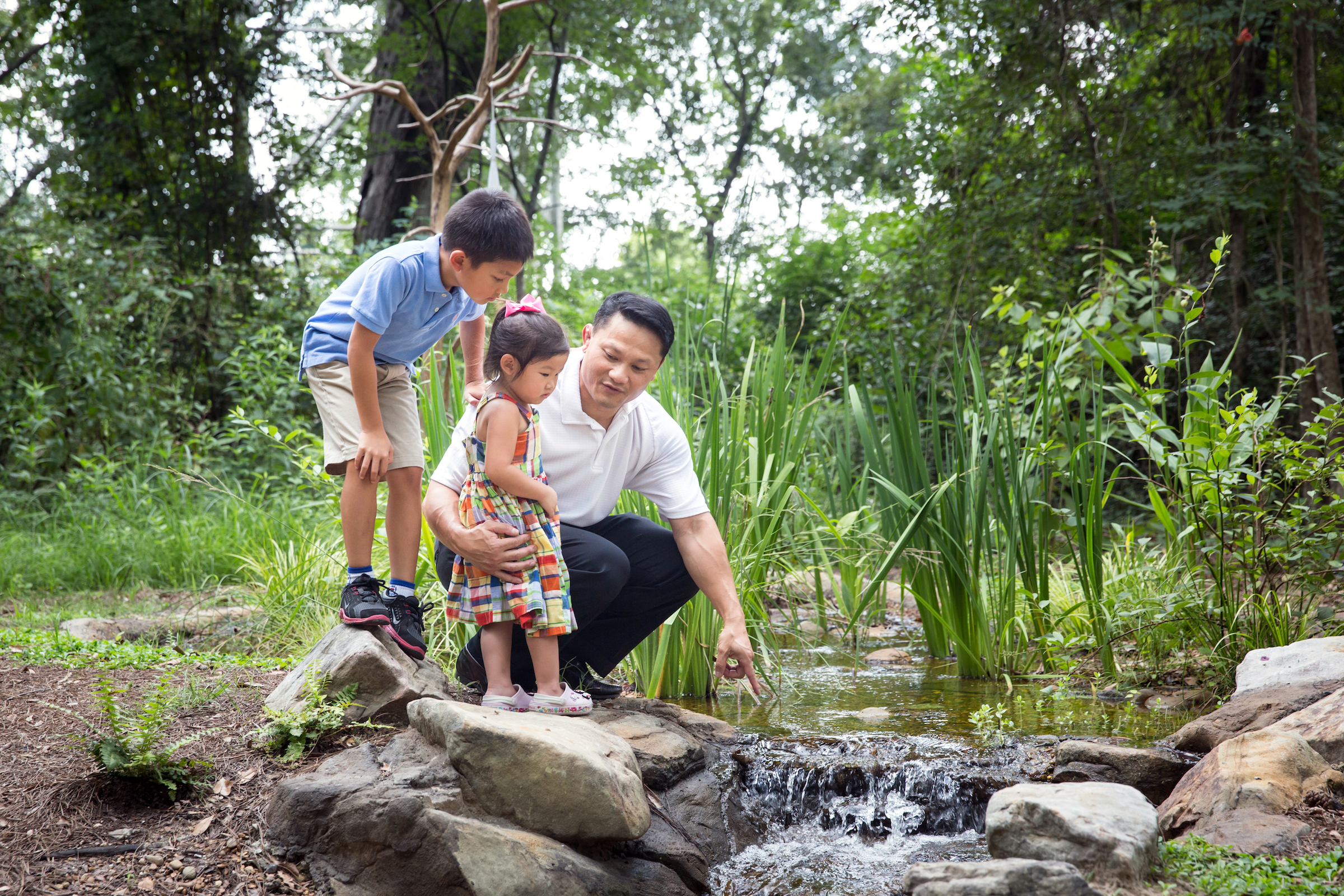 A father and two children sit by a creek. The dad is pointing towards the water.