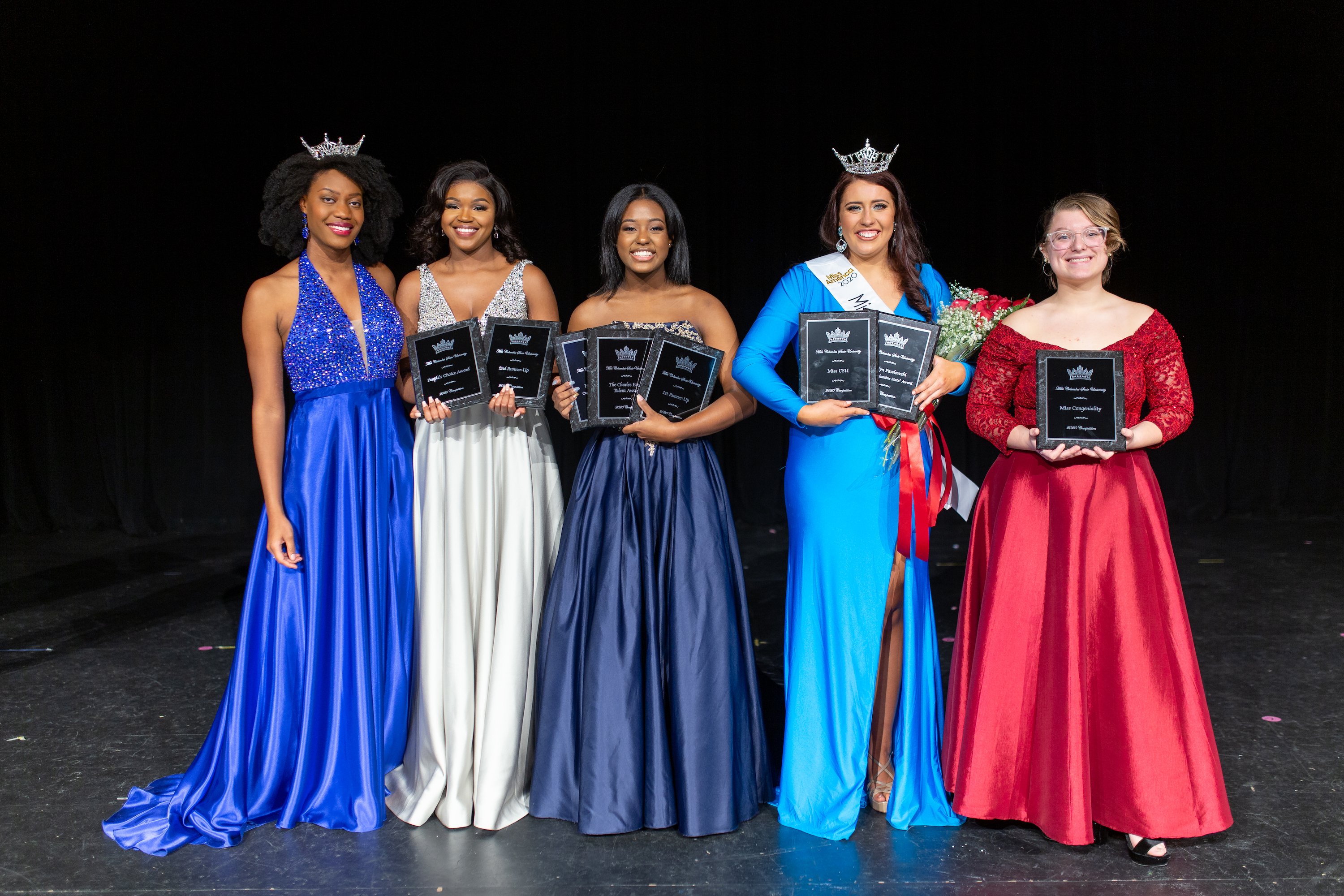 Five women in evening gowns are wearing tiaras and holding awards.