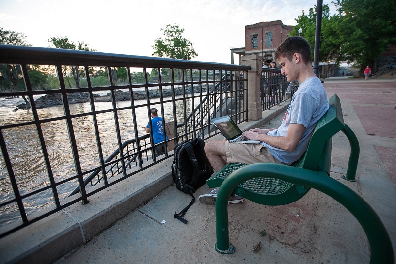 A student is sitting on a bench by a river walk working on his laptop