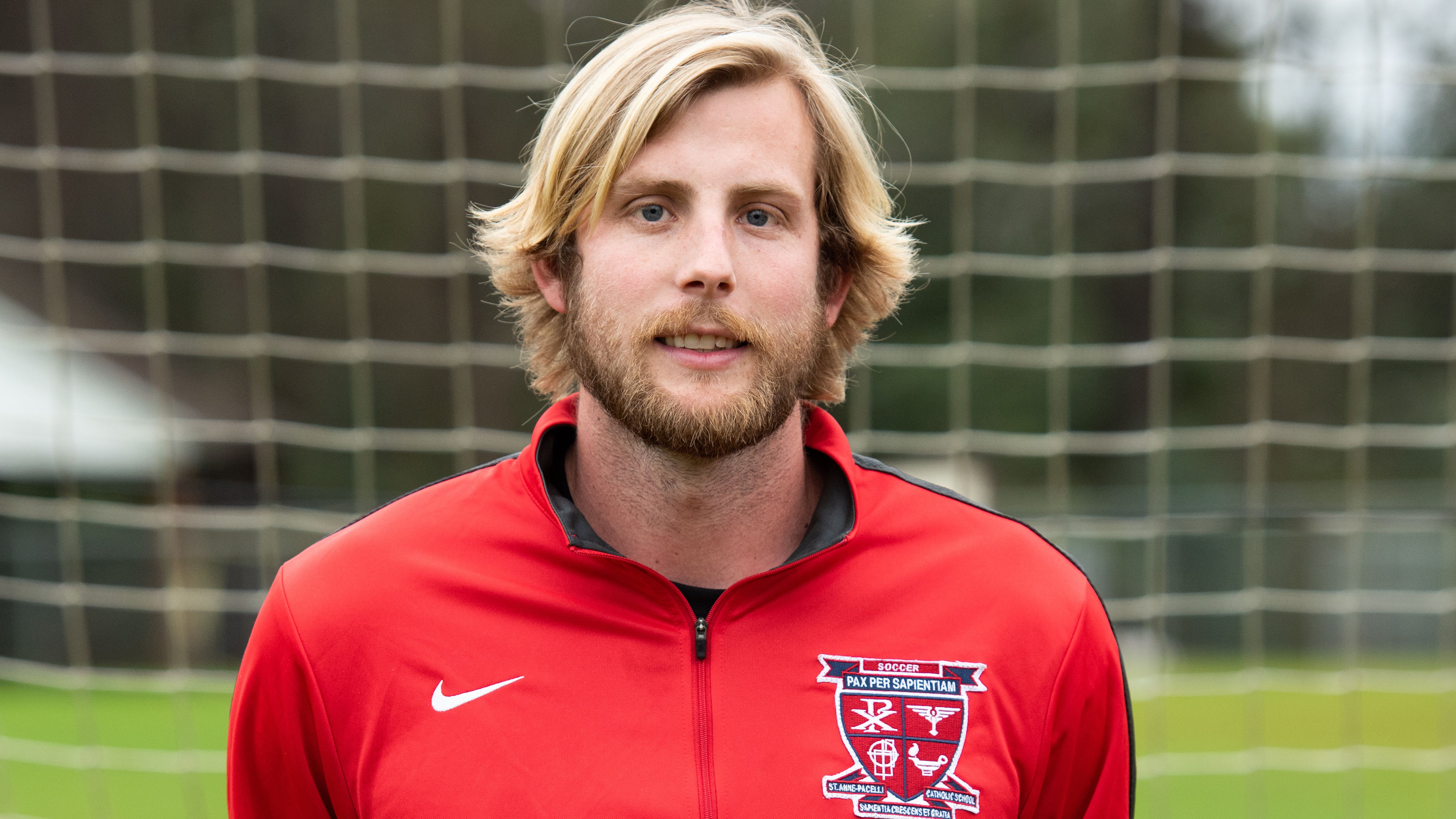 Zach Corcoran headshot in red sweatshirt in front of soccer goal.